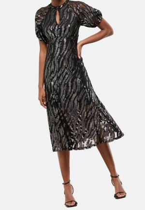 Femme portant une robe midi noire avec des manches bouffantes et un motif zèbre en sequins, associée à des sandales noires à talons hauts et bride à la cheville.