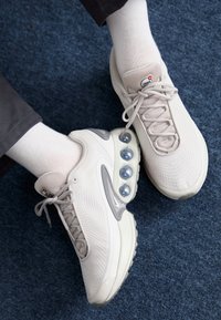 Sneaker grigie chiare con tomaia in mesh, accenti in gomma rotondi e pod di ammortizzazione ad aria visibili nella suola, abbinate a calze bianche.