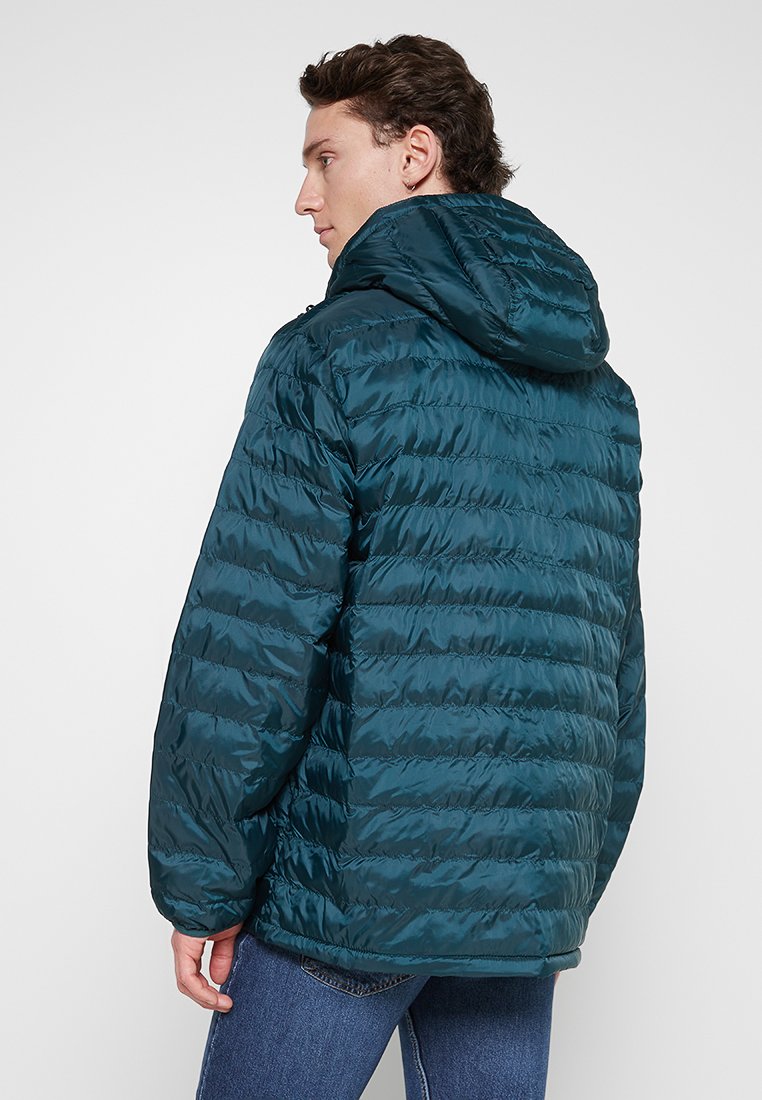 Man die zijwaarts kijkt in een donkerblauwe gewatteerde jas met capuchon en blauwe jeans tegen een effen lichte achtergrond.