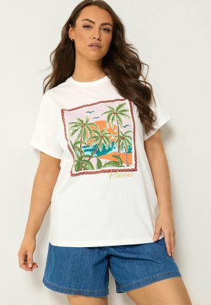Kvinde iført hvid T-shirt med farverigt Hawaii palmetræ-tryk og blå denimshorts, poserer mod en neutral baggrund.