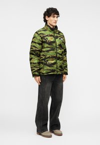 Μπουφάν τύπου puffer με πράσινο και μαύρο φωσφοριζέ μοτίβο παραλλαγής, με ψηλό κολάρο, κλείσιμο με φερμουάρ και λογότυπο. Συνδυασμένο με μαύρο τζιν φαρδιάς γραμμής.