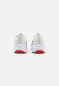 Weiße Sportschuhe mit glatter Textur, die rote Akzente an der Ferse und an der Seite aufweisen, sowie eine gummierte Sohle mit einer leichten Krümmung.