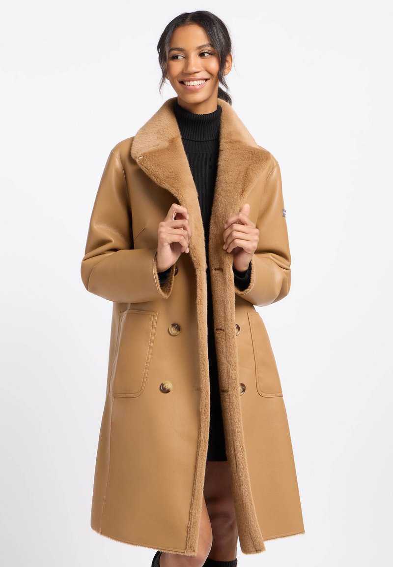 Camelkleurige jas met een nepbont kraag, dubbelgeknoopte knopen en twee voorkezen. Glad textuur en knielange ontwerp.