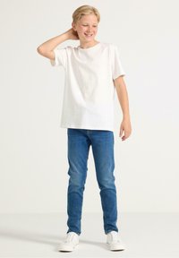 T-shirt in cotone bianco, vestibilità rilassata, abbinata a jeans in denim blu. Calzature: sneakers bianche. Il modello si trova in una postura casual con una mano sul collo.
