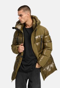 Olijfgroene pufferjacket met een glanzende afwerking, voorzien van een capuchon, voorzakken met rits en zwarte koorden, gedragen over een zwart shirt.