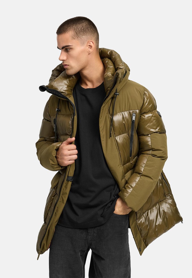 Olijfgroene pufferjacket met een glanzende afwerking, voorzien van een capuchon, voorzakken met rits en zwarte koorden, gedragen over een zwart shirt.