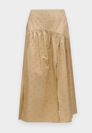 Kobieta z długimi włosami nosi beżową krótką kurtkę ze złotymi guzikami, biały bezrękawnik, szeroką brązową spódnicę z małymi wzorami i czarne szpilki.