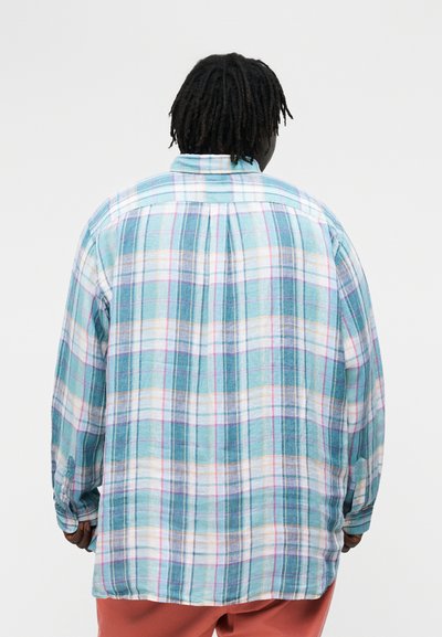 Osoba s krátkymi dredmi, oblečená v voľnej kockovanej košeli s dlhým rukávom v tyrkysovej a bielej farbe, stojaca chrbtom k nám na jednoduchom pozadí.