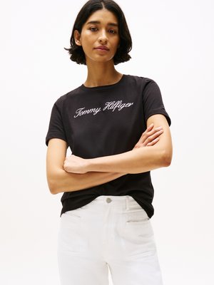 Černé bavlněné tričko s bílou výšivkou loga "Tommy Hilfiger", kulatým výstřihem a krátkými rukávy, v kombinaci s bílými džíny.