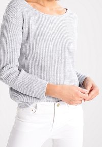 Světle šedý pletený svetr s širokým výstřihem, dlouhými rukávy a žebrovanou strukturou. Kombinovaný s bílými džíny se zlatými detaily.