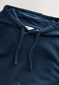 Tumesinine kapuutsiga dressipluus paeladega, nähtava kaeluse õmblusega ja väikese valge riidemärgisega kapuutsis sees.