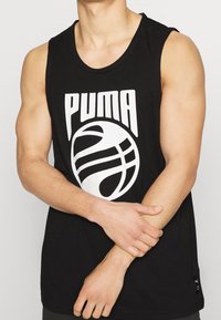 Homme portant un débardeur noir Puma avec un graphisme de basket-ball blanc, tenant son avant-bras gauche avec sa main droite.