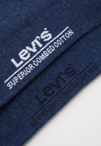 Tamsiai mėlyna medžiaga su balta ir tamsiai mėlyna tekstu. Pavaizduotas "Levi's" logotipas ir "superior combed cotton" kontrastingais šriftais. Minkšta tekstūra.