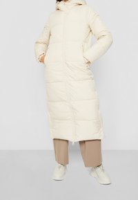 Beige lang pufferjakke med hætte, lynlåslukning, sidelommer og quiltet tekstur, parret med beige brede bukser og lette sneakers.