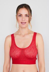 Czerwony bralet wykonany z teksturowanej tkaniny z falistymi wzorami, wyposażony w szeroką elastyczną taśmę z wytłoczonym logo oraz szerokie ramiączka.