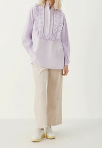 Blusa viola chiaro con dettagli in pizzo arricciato e pattina con bottoni, abbinata a pantaloni beige in corduroy a gamba larga e sneakers bianche.