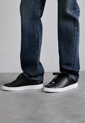 Sneaker nere in pelle con suole in gomma bianche, dotate di lacci neri, un accento con logo dorato e indossate con jeans blu scuro.