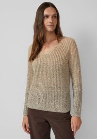 Vrouw in een beige en grijze gebreide V-hals trui en een bruine broek, staand tegen een effen grijze achtergrond.