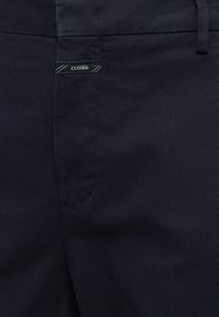 Pantalon bleu marine foncé montrant la poche avant et une petite étiquette noire cousue avec le mot "CLOSED" en fil argenté.