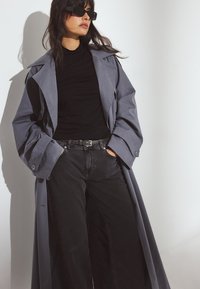 Grijze trenchcoat met brede revers, gecombineerd met een zwarte coltrui en donkere jeans. De jas heeft een soepele textuur en een oversized pasvorm.