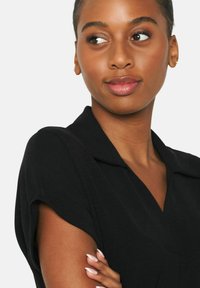 Camicia nera con colletto, maniche corte e arrotolate. Tessuto morbido e design aderente. Il modello ha un'espressione dolce e trucco neutro.