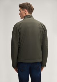 Giubbotto imbottito leggero di un verde scuro, con colletto alto e polsini abbottonati, indossato sopra jeans blu, che mette in mostra un design pulito e minimale.