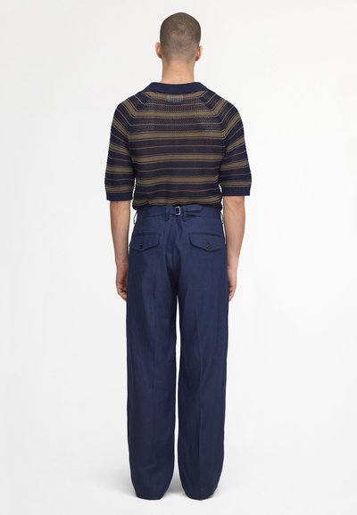 Homme aux cheveux courts portant un pull à rayures bleu marine et olive et un pantalon large taille haute bleu marine, debout de dos.