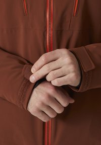 Hnědá bunda s oranžovým zipem; má nastavitelné manžety se suchým zipem. Detailní záběr na ruce, které upravují manžetu.