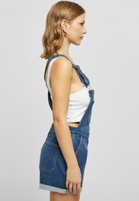 Denim overalls with a fitted shape, featuring adjustable shoulder straps, front pocket, and rolled hem shorts. Worn over a white crop top.