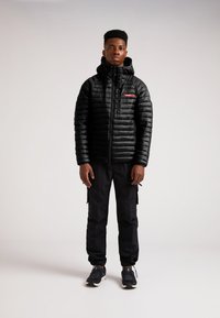 Giacca nera isolante con zip frontale e cappuccio, dotata di una toppa con logo rosso. Indossata con pantaloni cargo neri e scarpe sportive.