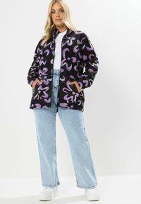Czarna polarowa kurtka z fioletowym wzorem kwiatowym, na zamek z przodu i bocznymi kieszeniami, zestawiona z jasnoniebieskimi dżinsami i białymi sneakersami.