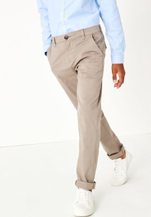 Homme portant un pantalon beige retroussé, une chemise bleu clair et des baskets blanches marchant devant un fond blanc uni.
