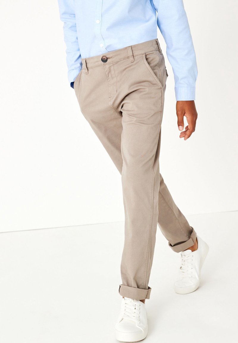 Homme portant un pantalon beige retroussé, une chemise bleu clair et des baskets blanches marchant devant un fond blanc uni.