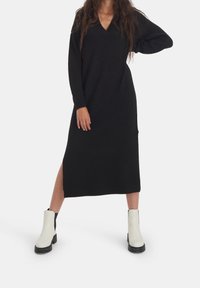 Svart långärmad klänning med V-ringning och sidenslits, tillverkad av mjukt material. Matchad med vita och svarta ankelstövlar med tjock sula.