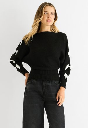 Black knitted sweater with wide sleeves, featuring white decorative bows at the cuffs and a fitted ribbed waistband. Paired with black jeans.