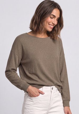 Femme aux cheveux bruns mi-longs souriante, portant un haut marron ample à manches longues et un pantalon blanc, une main dans la poche.