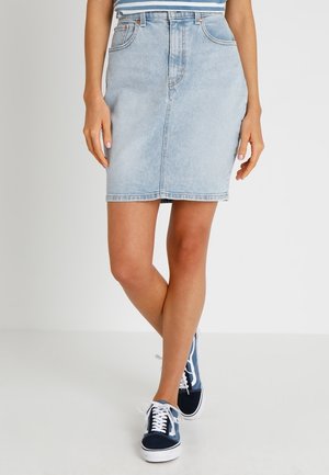 Woman wearing a light blue denim skirt and blue sneakers, standing with legs crossed against a plain white background.