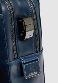 Primer plano de un bolso de cuero azul con cremallera metálica y hebilla azul y plateada con marca en el bolsillo lateral.