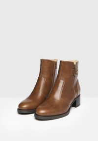Braune Leder-Stiefeletten mit abgerundetem Zehenbereich, seitlichem Reißverschluss und kleinem Blockabsatz. Glatte Textur mit kontrastierenden Nähten und einem dekorativen Riemen.
