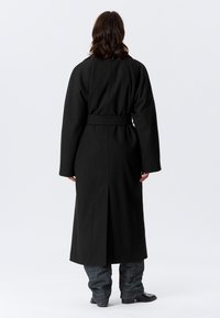 Čierny dlhý kabát z vlny, so zaväzovaním v páse, s širokými rukávmi a rozparkom vzadu pre lepšiu pohyblivosť.