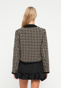 Svart og beige rutete tweedjakke med kort design, teksturert stoff og kontrasterende kant, parret med et svart volangskjørt.