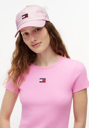 Ung kvinde med bølget hår iført en lyserød Tommy Jeans-kasket og en matchende lyserød ribstrikket t-shirt med logo på brystet.