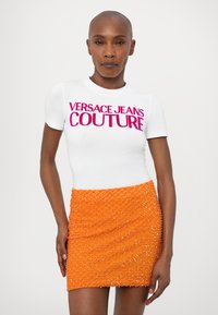 Fehér, testhezálló póló, élénk rózsaszín "VERSACE JEANS COUTURE" felirattal, melyet egy rövid, gyöngyszerű díszítéssel ellátott narancssárga szoknya kísér.