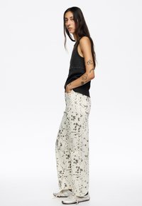 Top sin mangas negro con relieve y tachuelas plateadas, combinado con pantalones de pierna ancha color blanco roto con manchas grises. Llevado con zapatillas plateadas.