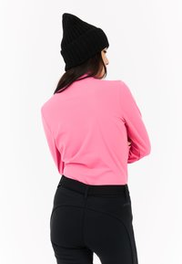 Różowa bluzka z długim rękawem i wysoką szyją, w zestawieniu z czarnymi dopasowanymi spodniami; model nosi czapkę z prążkowanej dzianiny w kolorze czarnym. Gładka tekstura materiału.