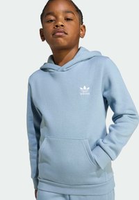 Svijetloplava hoodica s prednjim džepom, kapuljačom na vezanje i bijelim Adidas logotipom na prsima; izrađena od mekog, teksturiranog materijala.