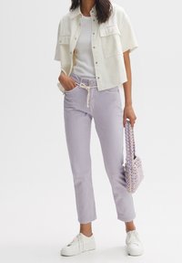 Veste blanche à manches courtes portée sur un haut blanc, associée à un pantalon lavande clair et des baskets blanches. Petit sac à main tissé violet en main.