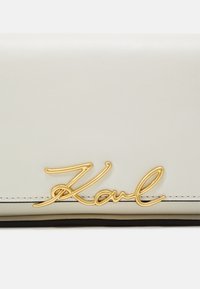 Portefeuille en cuir blanc avec un logo "Karl" en cursif doré, avec des coutures noires le long des bords. Texture lisse et design minimaliste.