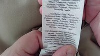 Étiquette de vêtement avec instructions d'entretien et matériaux, principalement en tissu blanc avec texte imprimé noir. Indique une teneur en polyester recyclé à 100%.