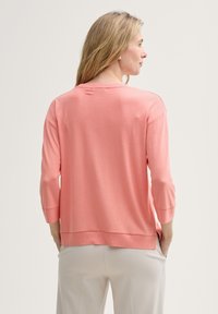 Maglione corallo con maniche a tre quarti, texture morbida, scollatura rotonda, vestibilità morbida e un orlo che scende leggermente sul retro.
